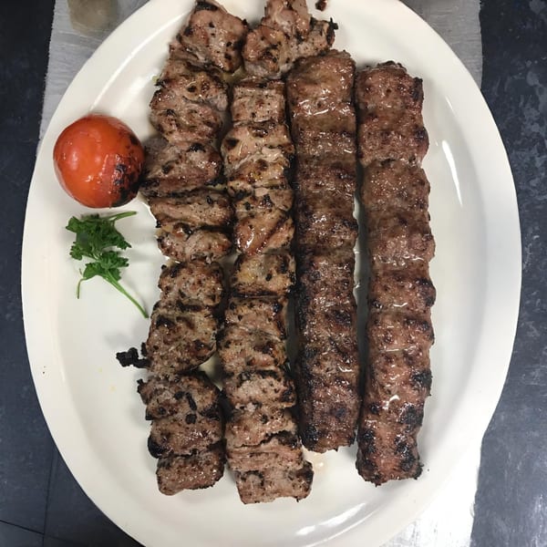 Grilled Mediterranean koobideh and shish kebab skewers with a roasted tomato and parsley on a white plate.