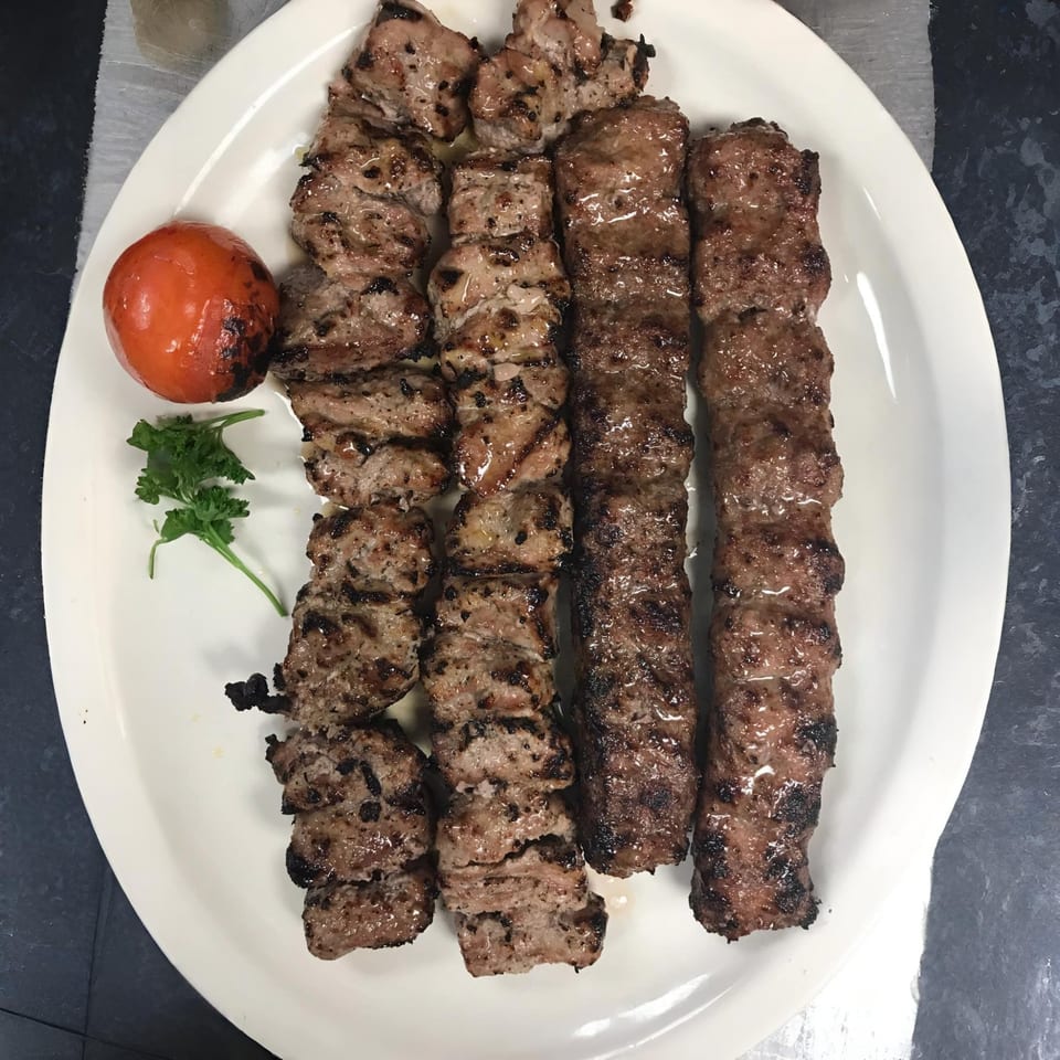 Grilled Mediterranean koobideh and shish kebab skewers with a roasted tomato and parsley on a white plate.